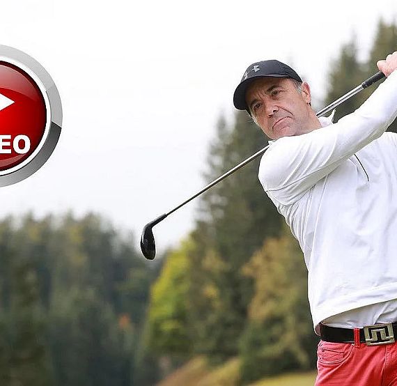 Ein Mann in weißem Pullover und roter Hose schwingt einen Golfschläger auf einem Golfplatz. Im Hintergrund sind Bäume und ein grauer Himmel zu sehen.