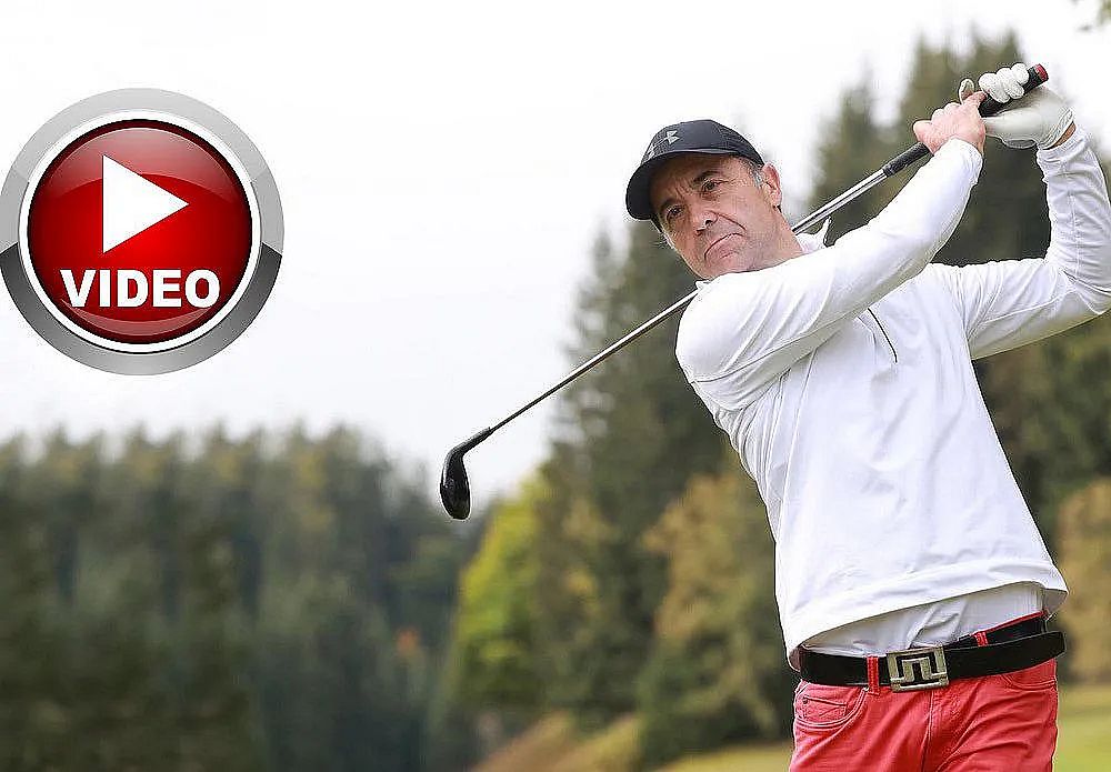 Ein Mann in weißem Pullover und roter Hose schwingt einen Golfschläger auf einem Golfplatz. Im Hintergrund sind Bäume und ein grauer Himmel zu sehen.