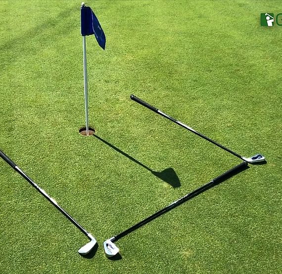 Three golf clubs form a square around a golf hole on a green. A blue flag is placed in the hole, and the clubs cast distinct shadows.