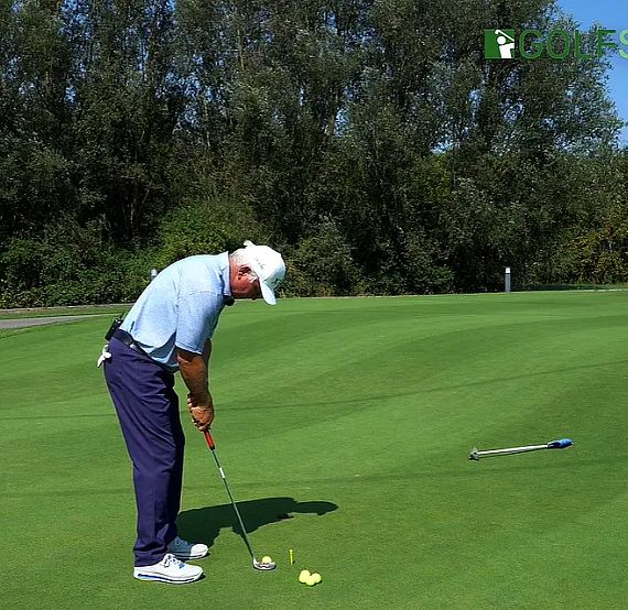 A golfer in a light blue shirt and dark pants practices putting on a green golf course, surrounded by trees, with a club and ball nearby.