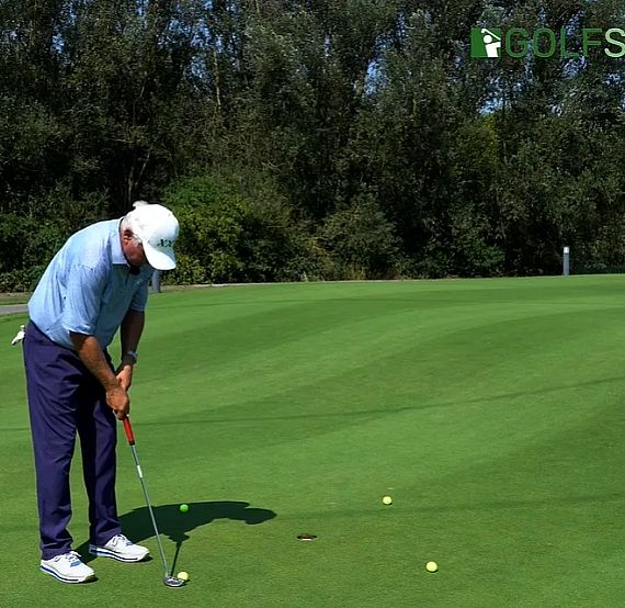 Golfer practicing putting on a green with trees in the background, wearing a blue shirt and dark pants.