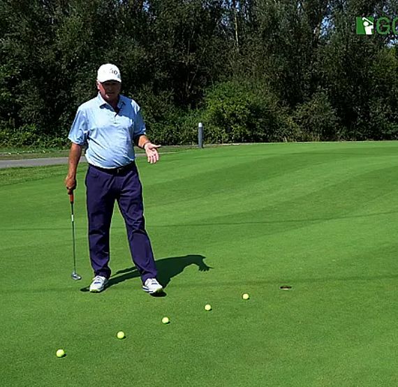 A golfer on a green field explaining putting. He's holding a putter with several balls lined up. Trees and a clear sky in the background.