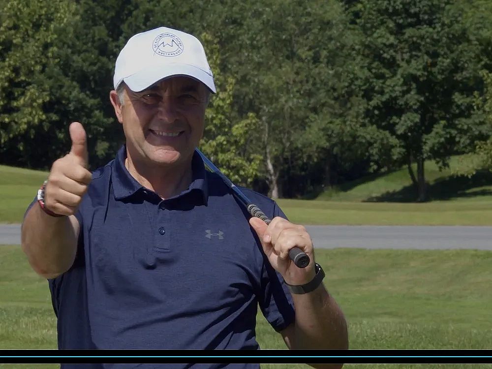 Person in navy shirt and white cap gives thumbs up on a golf course, holding a club, with green trees behind.