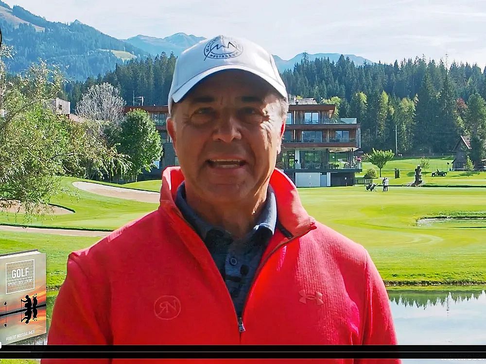 Ein Mann in rotem Pullover und weißer Mütze steht auf einem Golfplatz, umgeben von grünen Bäumen und Bergen im Hintergrund bei klarem Himmel.