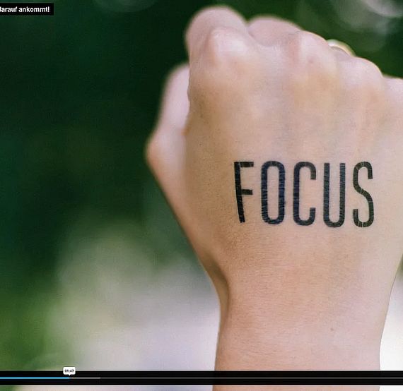 A close-up of a hand with 'FOCUS' tattooed on it. The background is blurred and green, indicating a natural setting, possibly trees or grass.