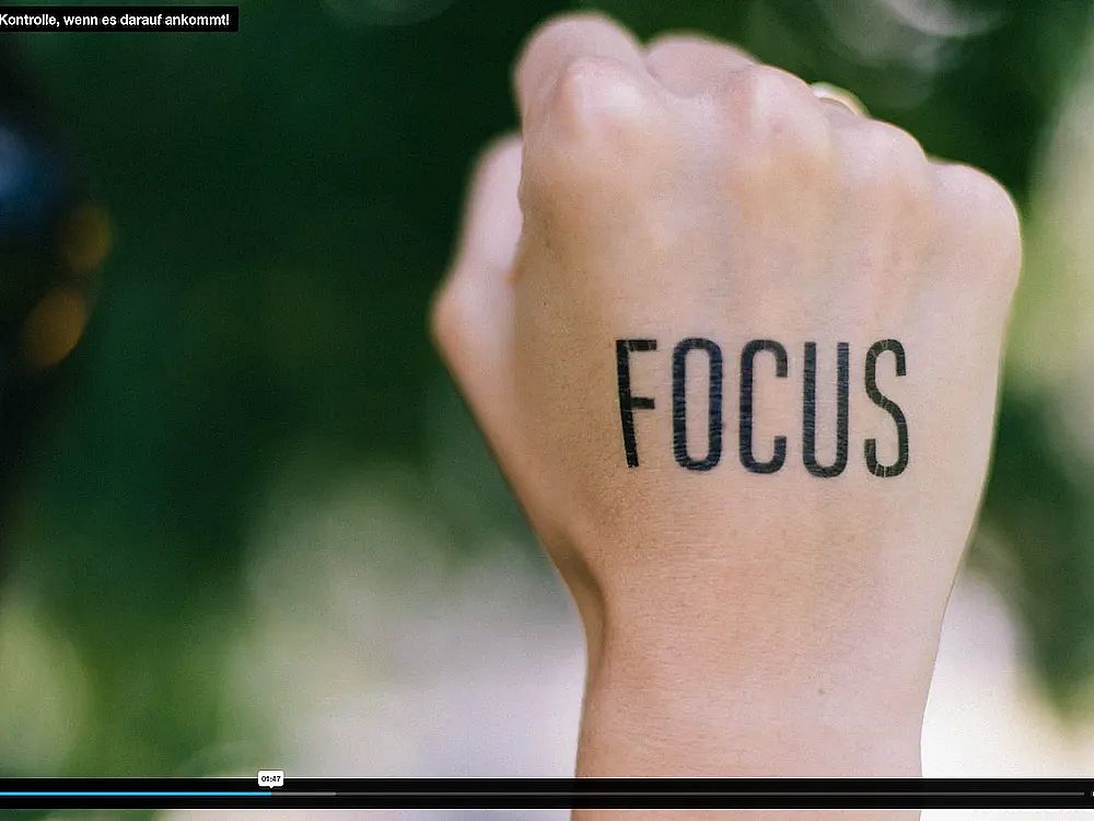 A close-up of a hand with 'FOCUS' tattooed on it. The background is blurred and green, indicating a natural setting, possibly trees or grass.