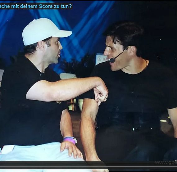 Zwei Männer sitzen auf einer Bühne. Einer trägt ein weißes T-Shirt und eine weiße Kappe, der andere ein schwarzes T-Shirt mit Headset. Sie diskutieren angeregt.