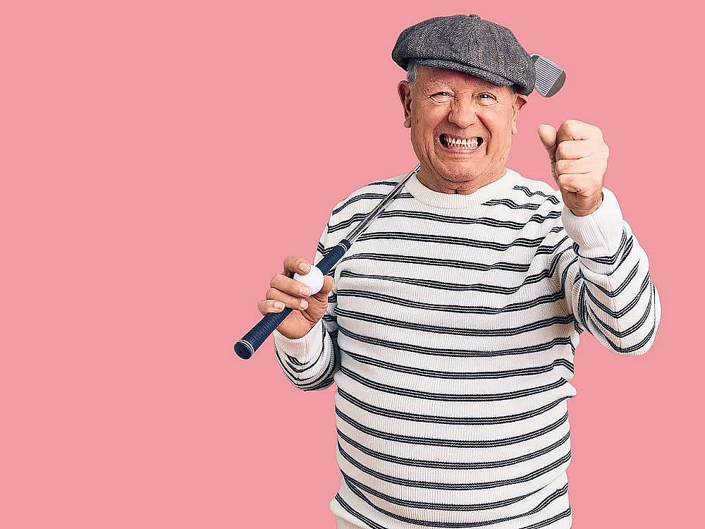 Elderly man in striped shirt and cap holding a paint roller, smiling with enthusiasm against a pink backdrop.