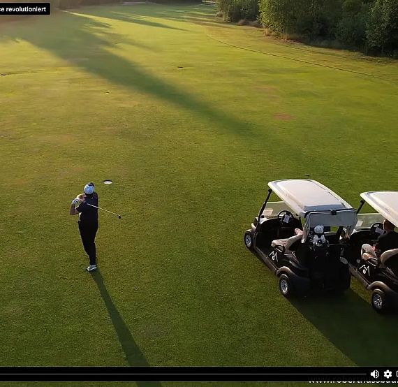 A golfer stands on a green field ready to swing, two golf carts nearby under a clear sky, infused with warm morning light.