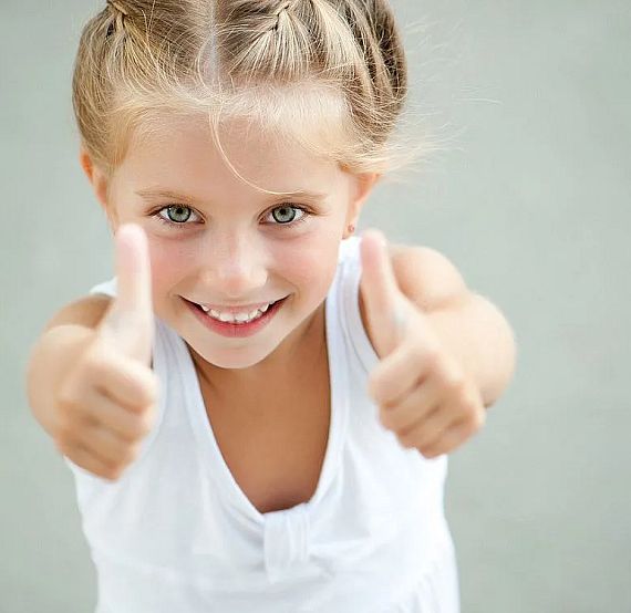 A smiling child with braided hair in a white top gives thumbs up. The background is gray, emphasizing the child's happy expression.