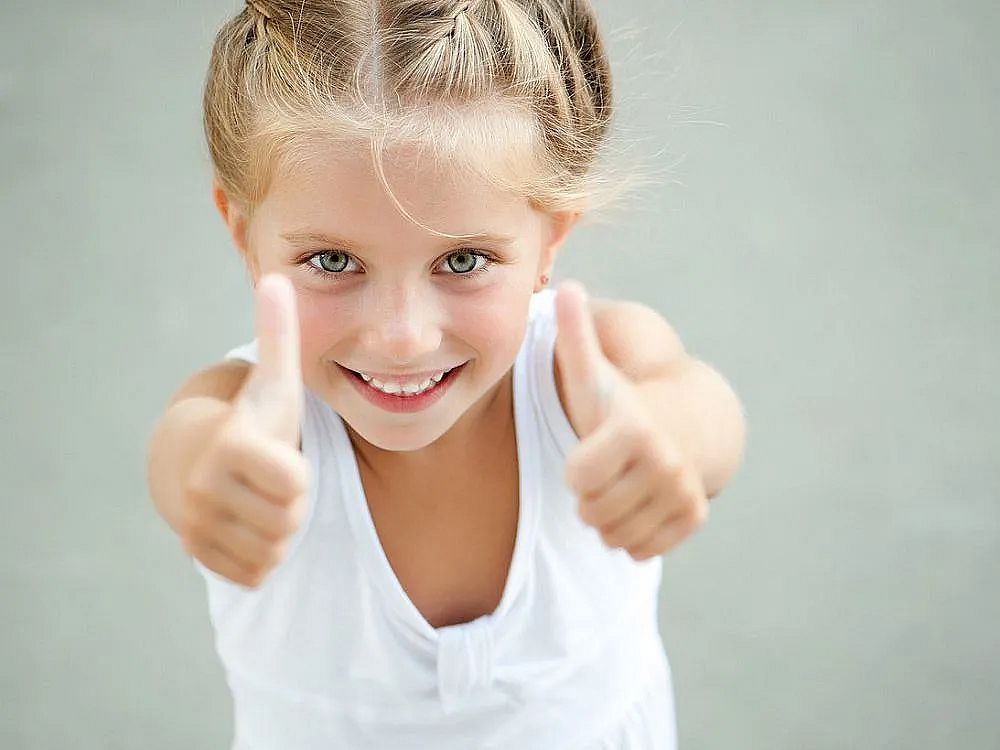 A smiling child with braided hair in a white top gives thumbs up. The background is gray, emphasizing the child's happy expression.
