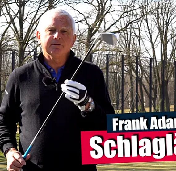 Man holding a golf club on a golf course, wearing a dark sweater and gloves, with trees in the background.