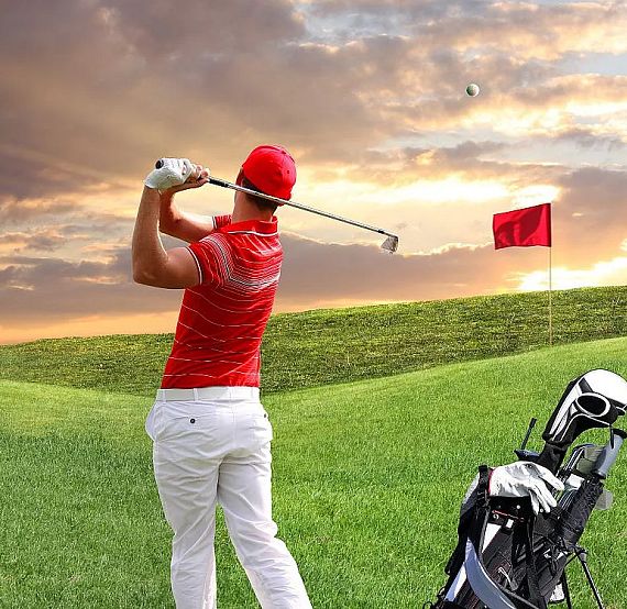 A golfer in red swings a club on a lush green course with a red flag, against a sky filled with dramatic sunset colors.