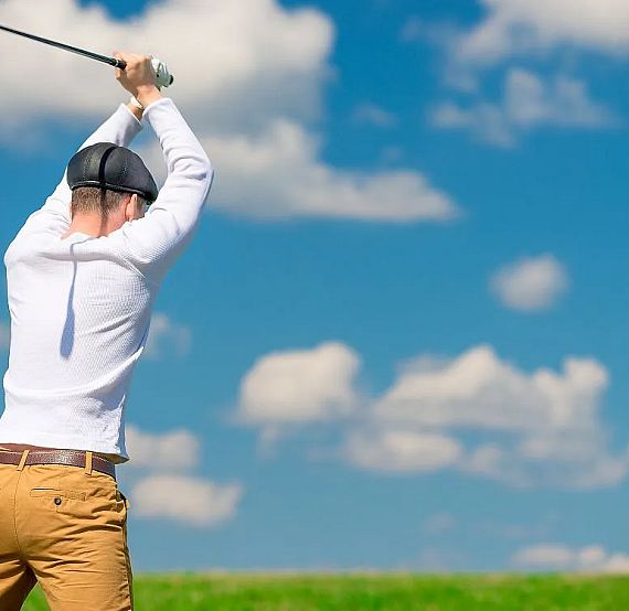 Man in a white shirt and cap swings a golf club on a sunny day, against a backdrop of blue sky with clouds and green grass.