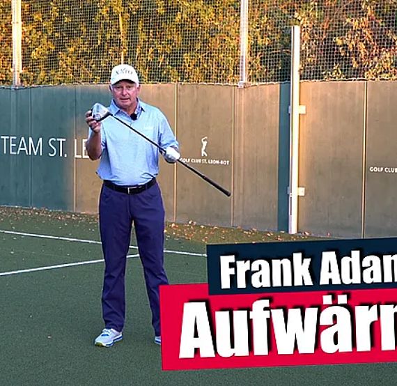 Golfer auf einem Trainingsfeld, hält einen Schläger, steht vor einem Zaun mit Beschilderung. Sonniger Tag, Herbstblätter im Hintergrund.