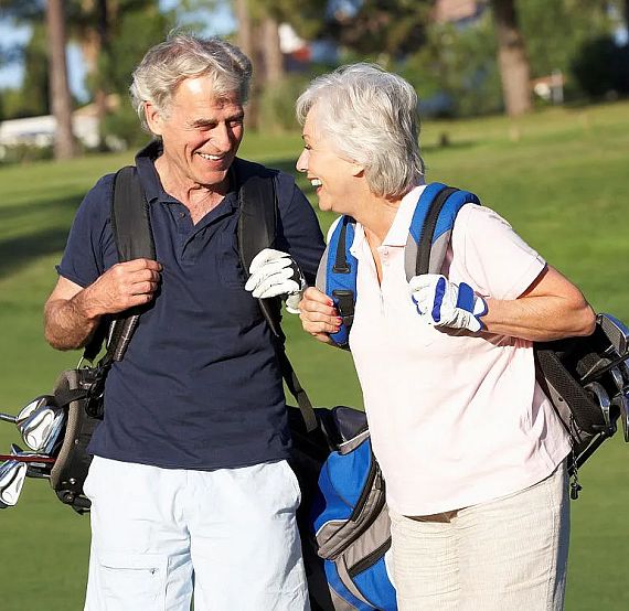 Senior couple smiling while carrying golf clubs on a sunny golf course, surrounded by greenery.
