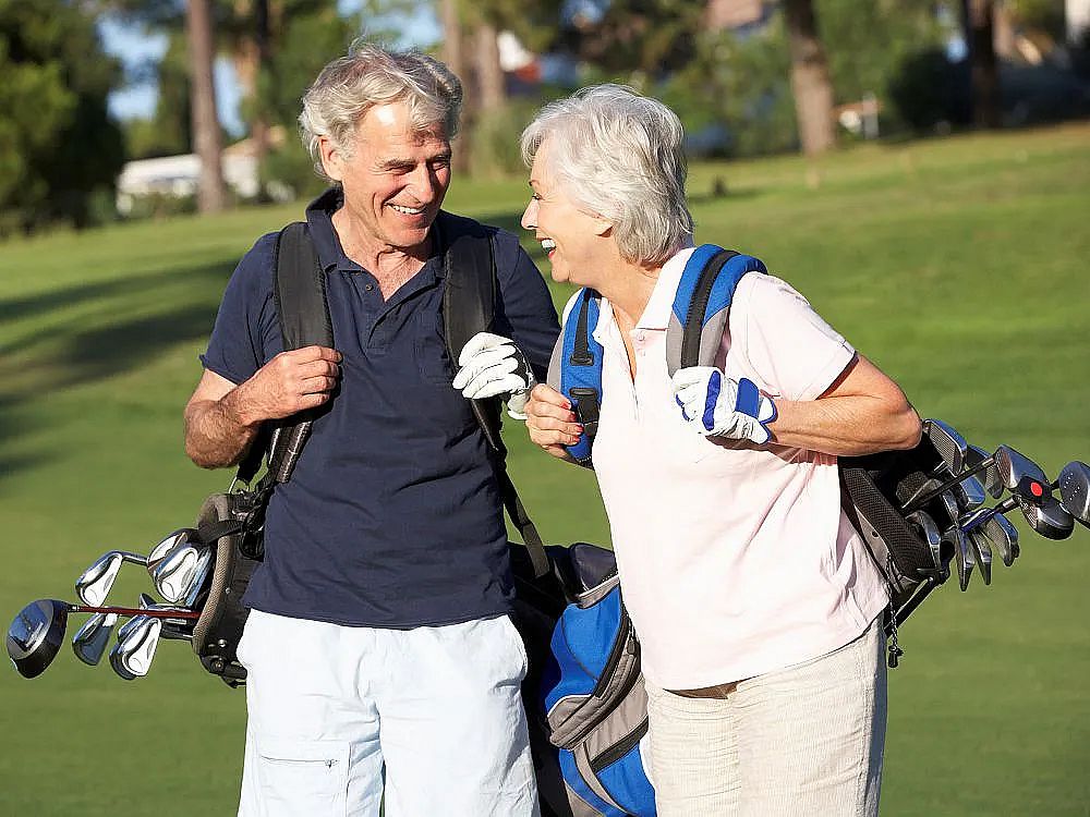 Senior couple smiling while carrying golf clubs on a sunny golf course, surrounded by greenery.