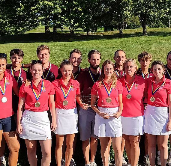 A group of men and women in matching red shirts and white skirts or shorts pose with medals against a backdrop of grass and trees under a blue sky.