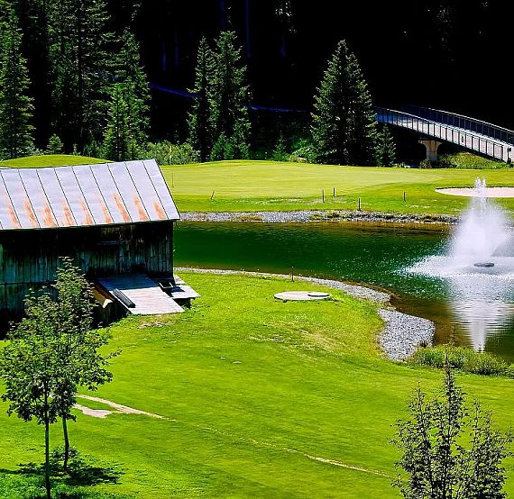 Scenic view of a golf course with a rustic cabin, a pond with fountain, and a bridge, set amidst green lawns and tall trees.
