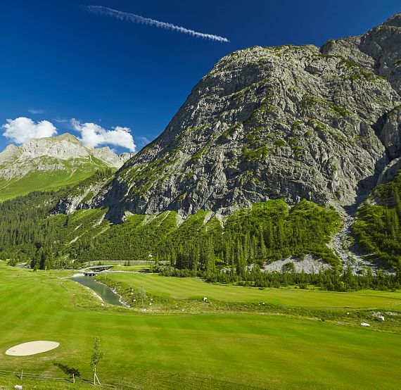 Golfplatz in alpiner Landschaft mit saftig grünem Rasen, umgeben von beeindruckenden Bergen und dichten Wäldern unter einem klaren blauen Himmel.