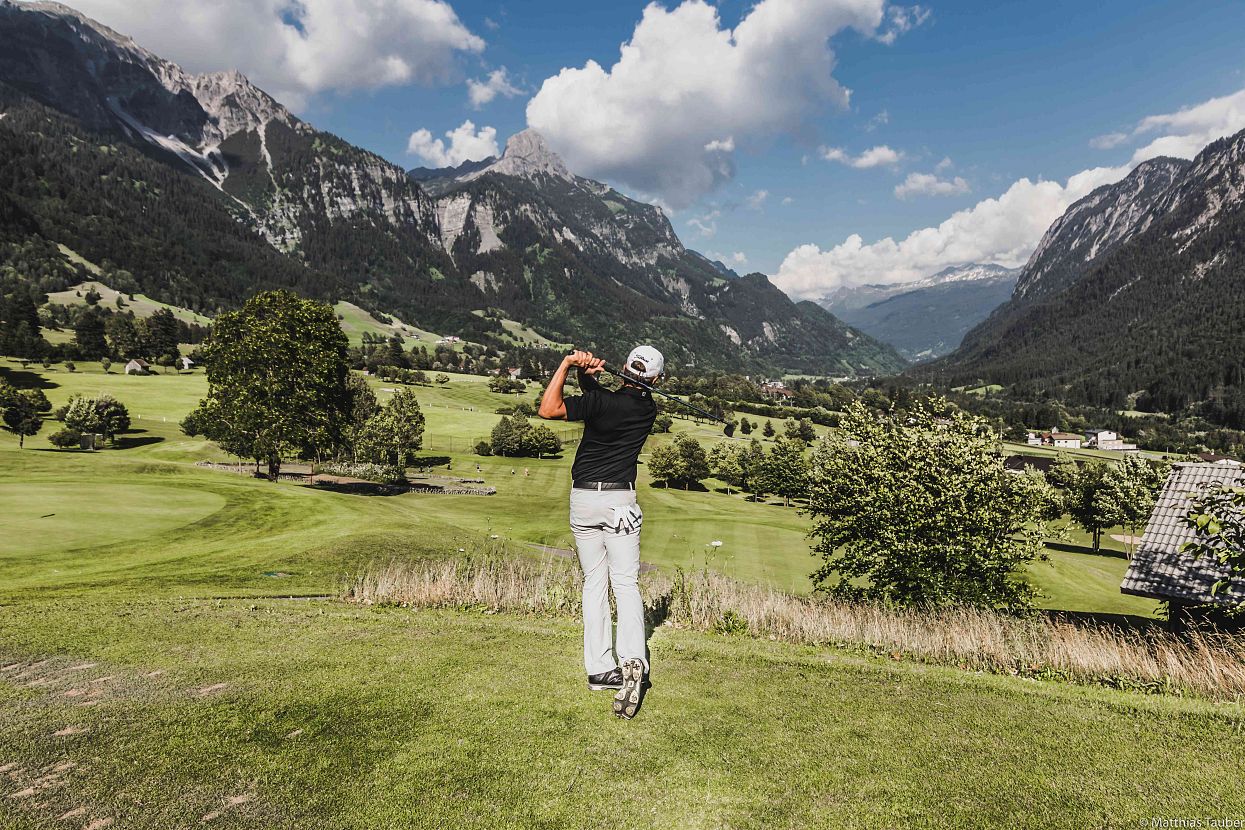 Golfer swings on a green course surrounded by mountains and a partly cloudy sky, creating a serene and picturesque setting.