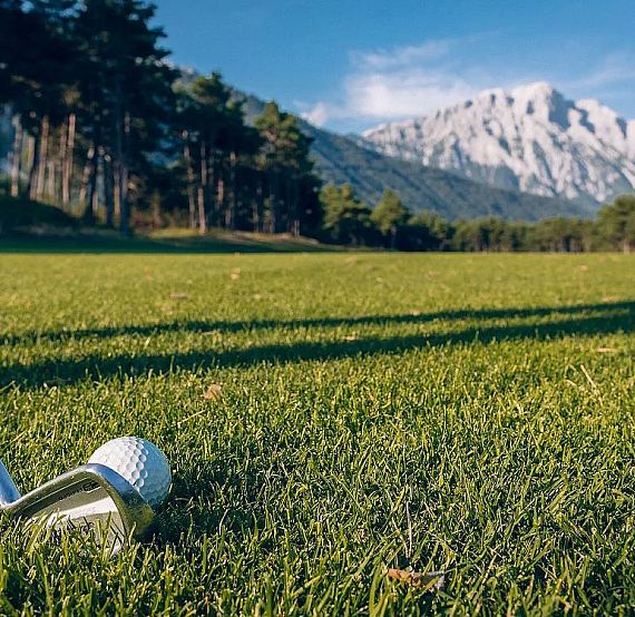Golf club and ball on green grass with pine trees and mountains in the background under a clear blue sky.
