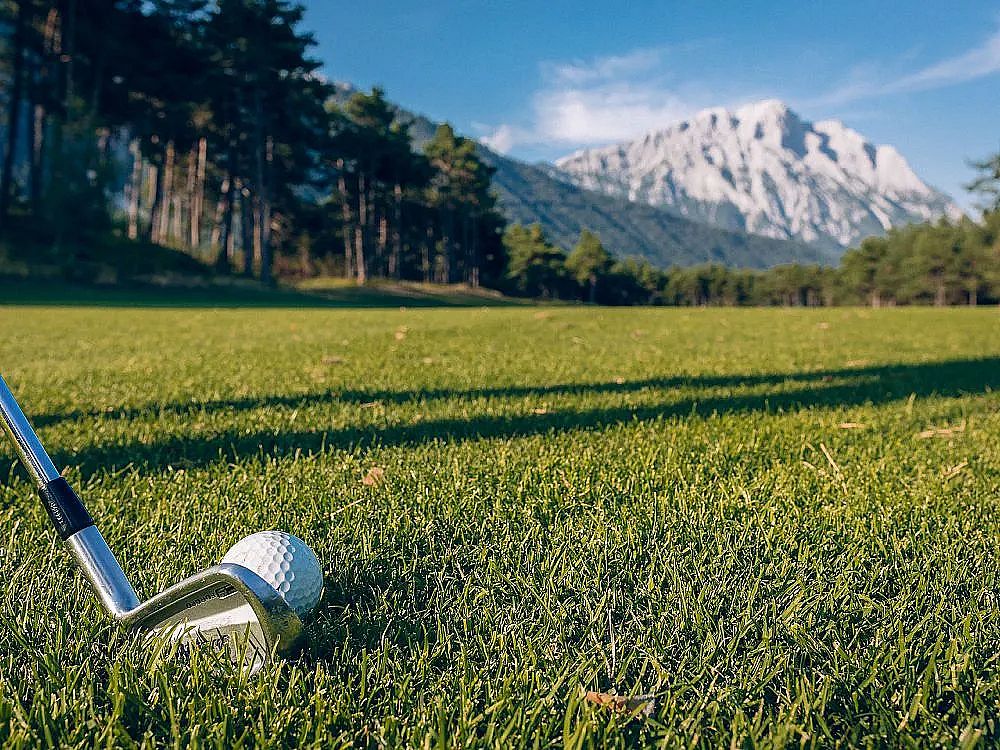 Golf club and ball on green grass with pine trees and mountains in the background under a clear blue sky.