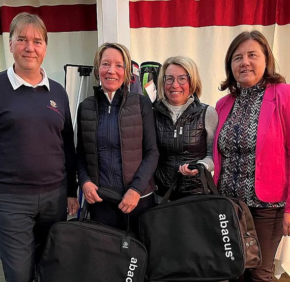 Four people pose together, smiling and holding branded bags. They appear to be at an event, against a backdrop of a red and white curtain.