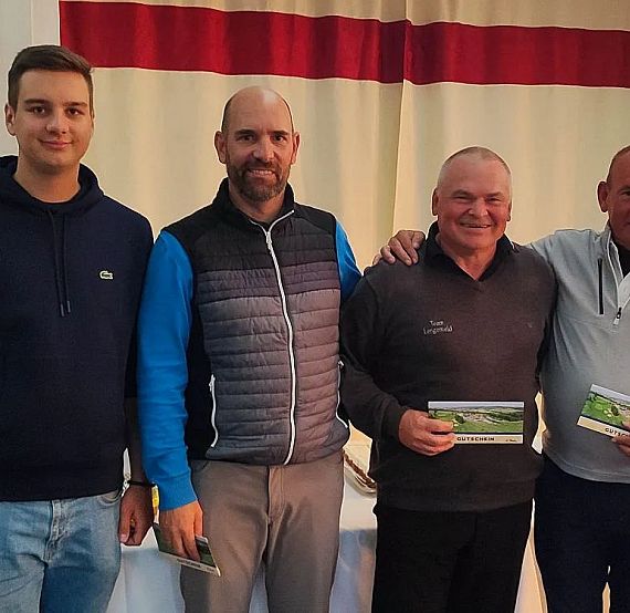 Vier Männer stehen lächelnd nebeneinander in einem Raum, zwei von ihnen halten Karten in den Händen. Sie sind lässig bis sportlich gekleidet.