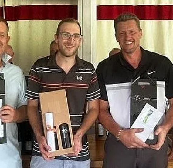 Four men standing together, each holding a gift box, smiling, in a casual setting with curtains in the background.