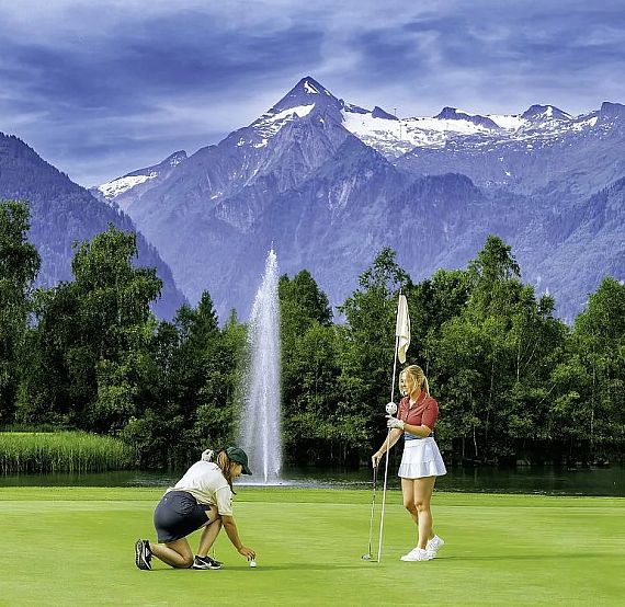 Zwei Personen auf einem Golfplatz, eine kniet und platziert einen Ball, die andere hält die Fahne. Im Hintergrund Berge und ein Springbrunnen.