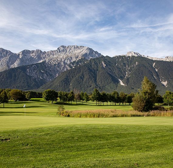 Grüne Golflandschaft mit gepflegtem Rasen, umgeben von Bäumen und Büschen, vor einer Kulisse aus beeindruckenden Bergen unter einem blauen Himmel.