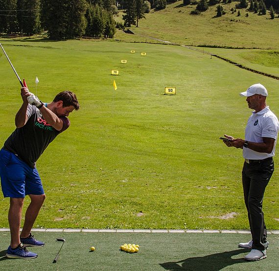 Ein Golfer bereitet sich auf einen Schlag auf einem grünen Fairway vor, während ein Trainer daneben steht, der Notizen auf einem Tablet macht.