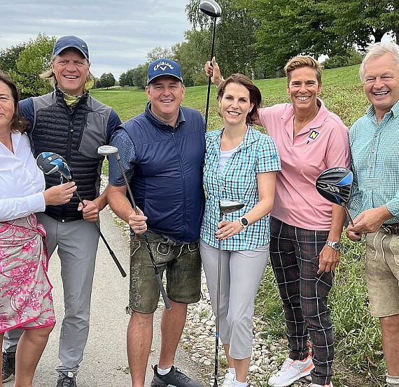 A group of six people standing on a pathway beside a grassy field, holding golf clubs and wearing casual golf attire.