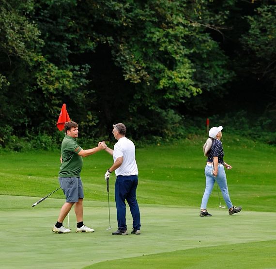 Two golfers shake hands on a green golf course, while another person walks nearby. Lush greenery and trees surround the scene.