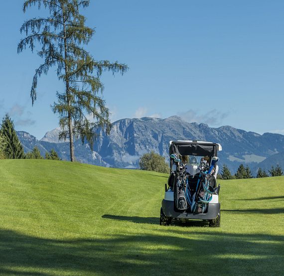 Golf cart on a green course with mountains in the background, trees around. Clear blue sky and tranquil setting.