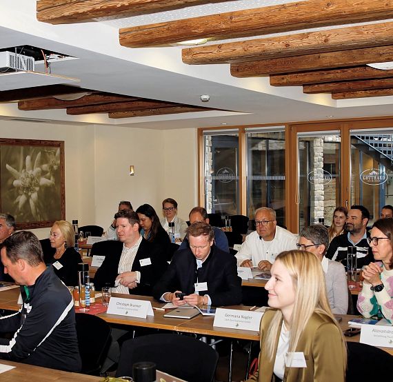 A group of people sit at long tables in a rustic room with wooden beams, focused on a presentation. Papers and drinks are on the tables, with large windows in the background.
