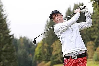 A golfer in a white shirt and red pants swings his club on a green course with trees in the background, hinting at a serene outdoor setting.
