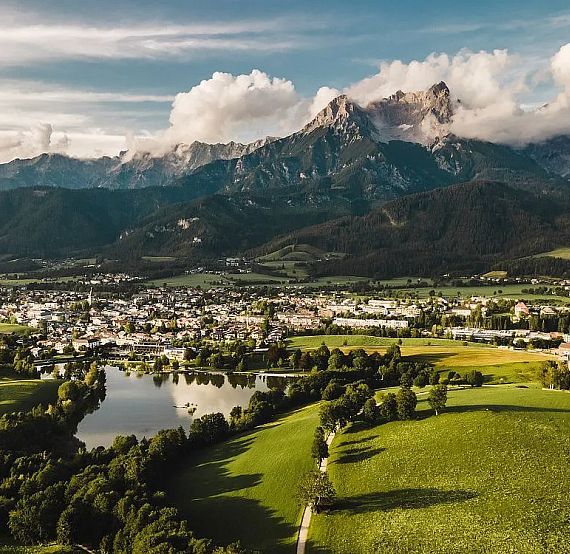 Aerial view of a lush valley with a lake, surrounded by mountains under a cloudy sky.