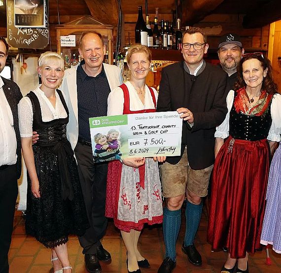 Eine Gruppe von Menschen in traditioneller bayerischer Tracht lächelt in die Kamera und hält einen großen Scheck in einem rustikalen Raum.