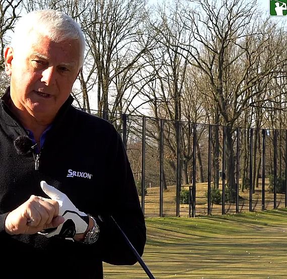 Man in dark jacket talks about golf outdoors with trees and golf course in background, on a sunny day, holding a golf glove with focused expression.