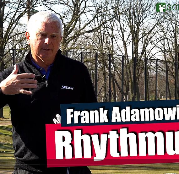 Man giving a golf lesson outdoors with trees in the background, holding a golf club, and a sign reading 'Rhythm' and 'Frank Adamowicz'.