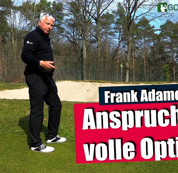 A man stands near a sand trap on a golf course, gesturing with trees in the background, under a blue sky.