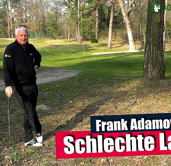 An elderly man in a black outfit stands with a golf club in a wooded area beside a golf course path.