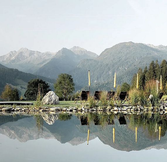 Eine malerische Berglandschaft mit einem ruhigen See im Vordergrund, umgeben von Liegestühlen und Sonnenschirmen. Im Hintergrund erheben sich majestätische Berge.