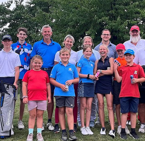 Gruppe von Menschen unterschiedlichen Alters in sportlicher Kleidung auf einem Rasen, posierend für ein Gruppenfoto. Im Hintergrund sind Bäume sichtbar.