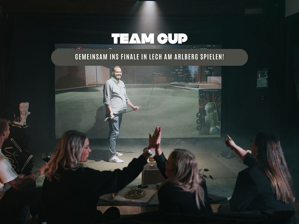 Gruppe von Menschen in einem dunklen Raum, die auf einen Bildschirm schauen, auf dem ein Spiel oder Präsentation gezeigt wird. Ein Mann steht vorne.