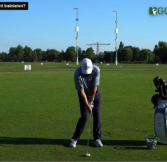 A golfer practices his swing on a green field with a golf bag and trees under a clear sky.