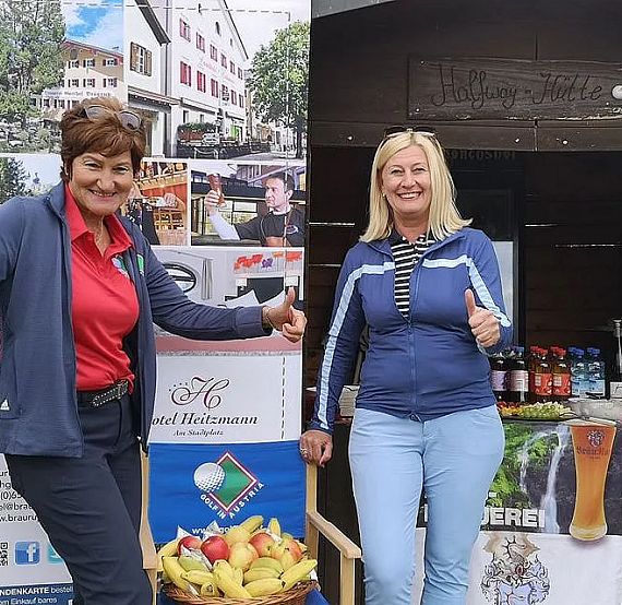 Two smiling individuals stand by an outdoor booth with promotional banners for hotels and beverages, giving thumbs up in a friendly gesture.