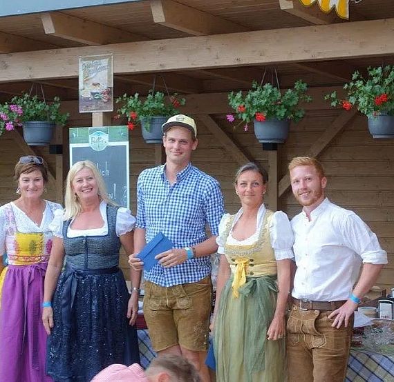Five people in traditional Bavarian clothing with dirndls and lederhosen, standing outside a wooden building decorated with hanging flower pots.